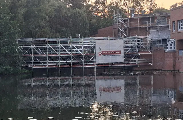 Mehrgeschossiges Brückengerüst aus Stahlrohren mit Arbeitsplattformen und VVV-Gerüstbau-Banner über einer Wasserfläche in Hamburg-Wilhelmsburg.